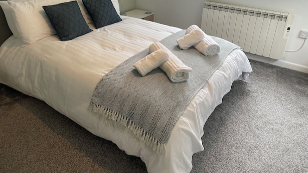 Een net opgemaakt bed met witte lakens, twee donkere kussens en een deken met een patroon.