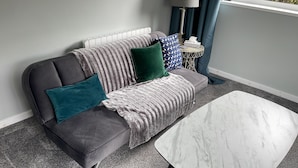 Een grijze bank met teal- en groen gekleurde kussens, een grijze deken en een witte marmeren salontafel in een kamer met grijs tapijt en een radiator.