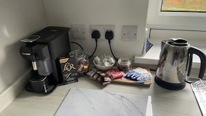 Een keukenblad met een koffieapparaat, een waterkoker, een pot koffiecups, en een snijplank met koffiecups en biscotti.