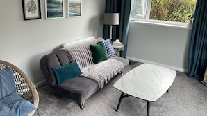 Een moderne woonkamer met een grijze bank, teal- en blauwe kussens, een witte salontafel en een rieten fauteuil.