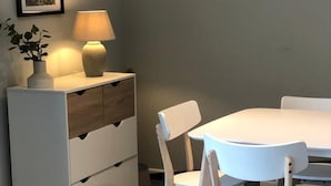 Een eethoek met een witte tafel en stoelen, een lamp op een bijzettafeltje en een lijst met een afbeelding aan de muur.