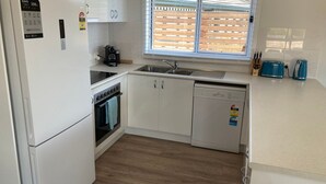 Een compacte keuken met witte kastjes, een inbouwoven, een vaatwasser en een raam met zonwering.