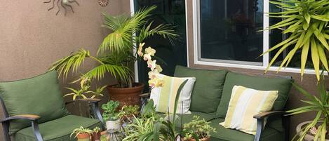 Un rincón al aire libre acogedor con sillas verdes acolchadas, una mesa de centro de madera con plantas en macetas y decoración en la pared.