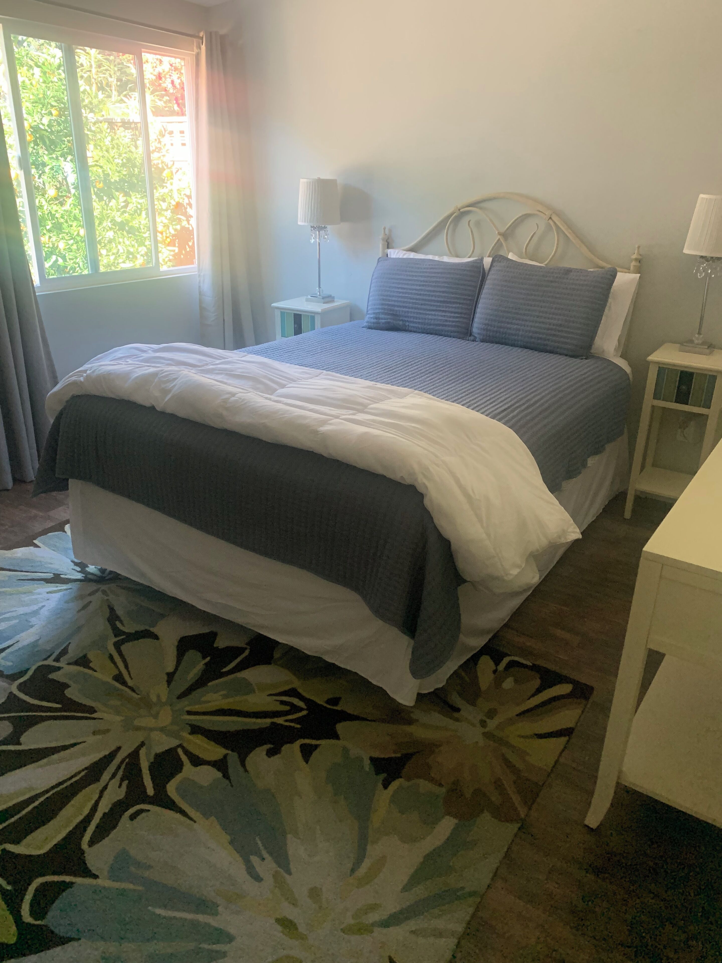 Een keurig opgemaakt bed met een wit-grijze deken, een blauw hoofdbord en een nachtkastje met een lamp.