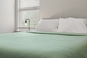 A bedroom with a bed, white pillows, a green blanket, and a window with blinds and a hanging light fixture. - Spacious & Comfortable 3 Bedroom Home with Garage (Fort Worth)