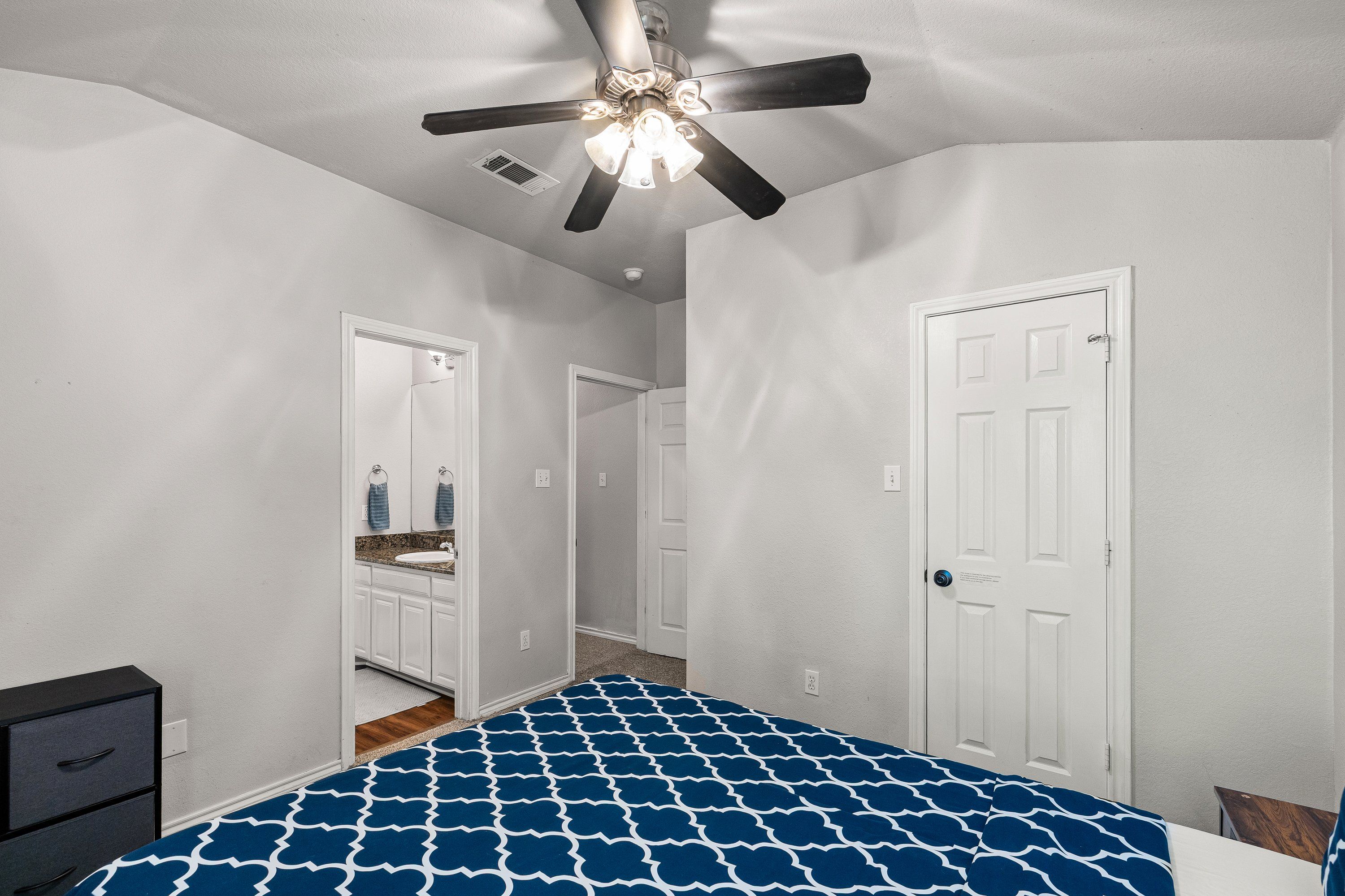 Un dormitorio con cama, ventilador de techo, una puerta blanca y baño visible a través de una puerta abierta.