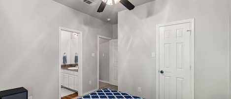 A bedroom with a bed, a ceiling fan, a white door, and a bathroom visible through an open door.
