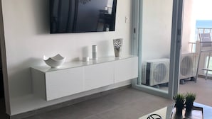 A modern living room with a flat-screen TV mounted on the wall, a white console table with decorative items, and a sliding glass door leading to a balcony with outdoor furniture.