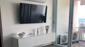 Un salon moderne avec un téléviseur à écran plat fixé au mur, une console blanche agrémentée d’objets décoratifs et une porte-fenêtre coulissante donnant sur un balcon équipé de mobilier d’extérieur.