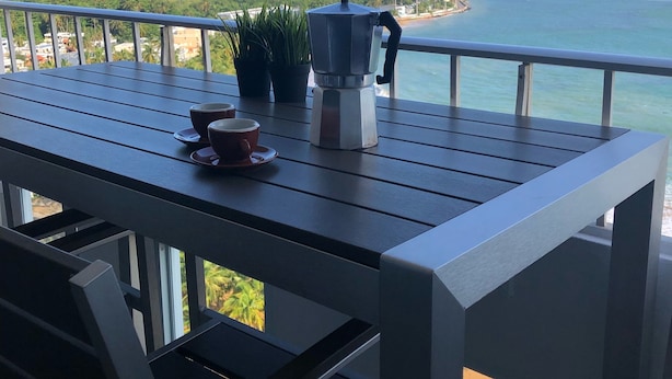 A balcony with a table, a coffee maker, and a cup. The view includes the ocean and a distant shoreline with buildings.