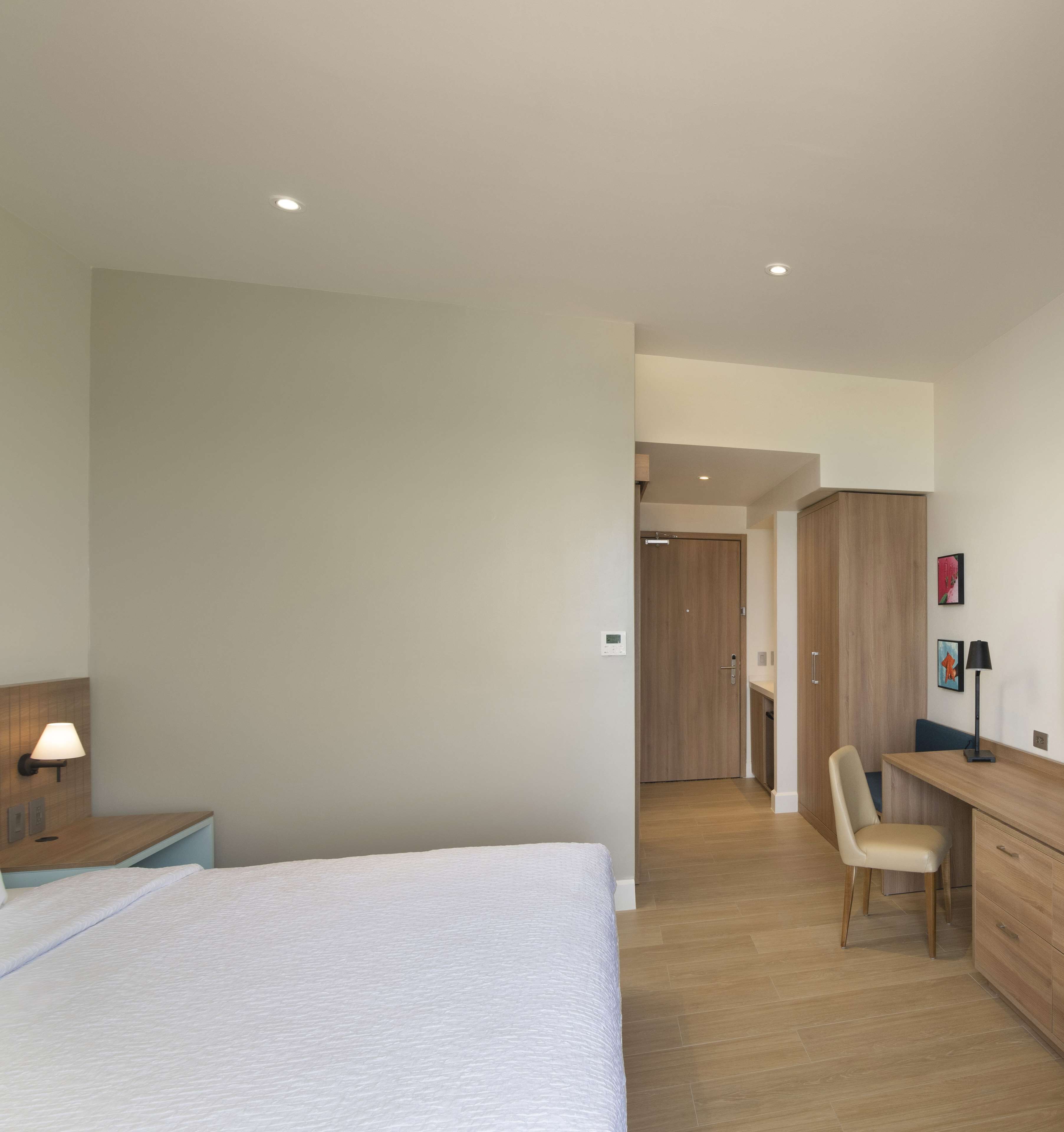 Een moderne slaapkamer met een groot bed, een bureau en een stoel. Er is een deur naar een badkamer en een kitchenette.