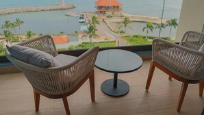 Een balkon met rieten stoelen en een kleine tafel, met uitzicht op een kustgebied met een pier en een gebouw met een rood dak.