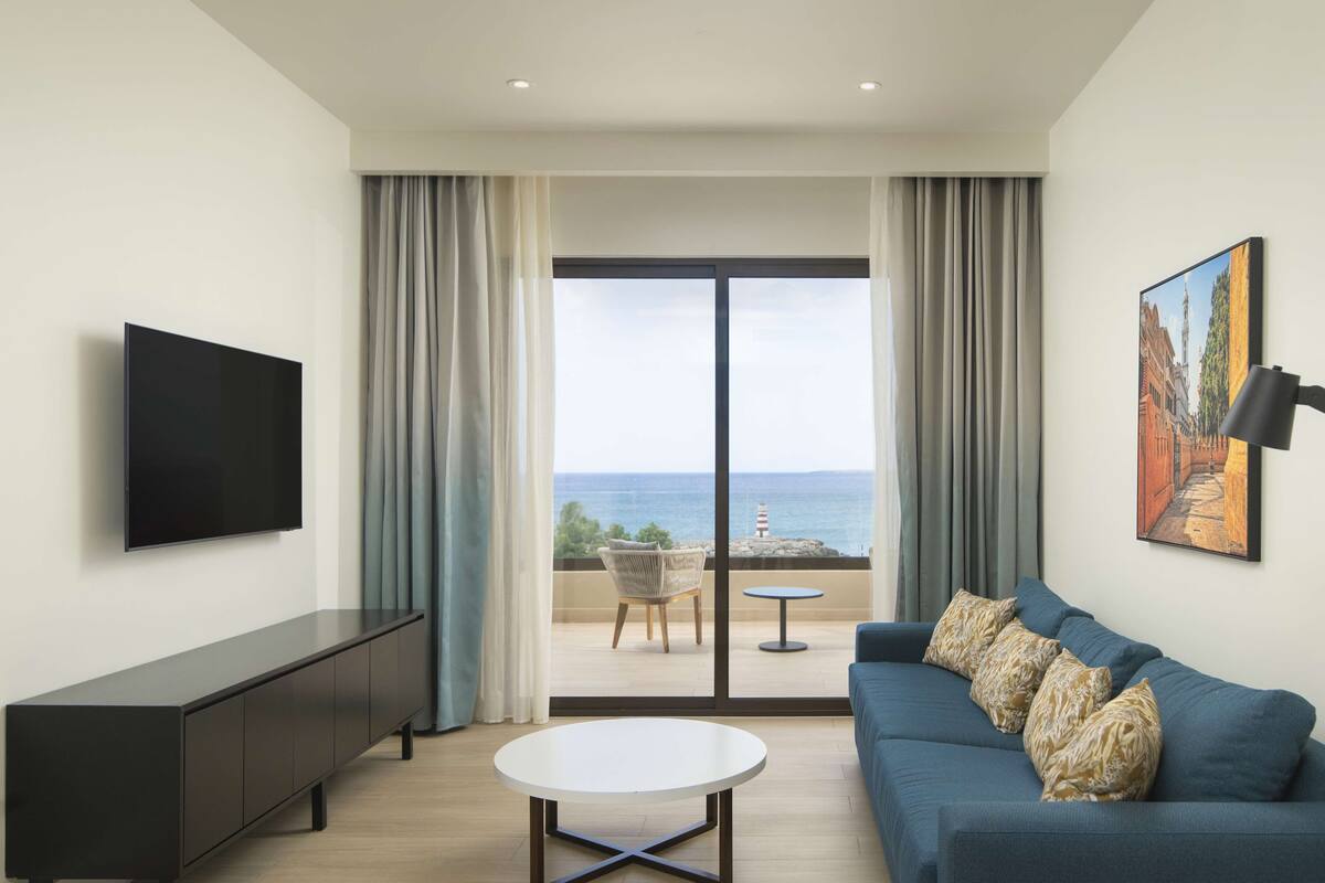 Un salon moderne doté d’un téléviseur à écran plat, d’un canapé agrémenté de coussins à motifs, d’une table basse ronde et d’un balcon offrant une vue sur la mer.