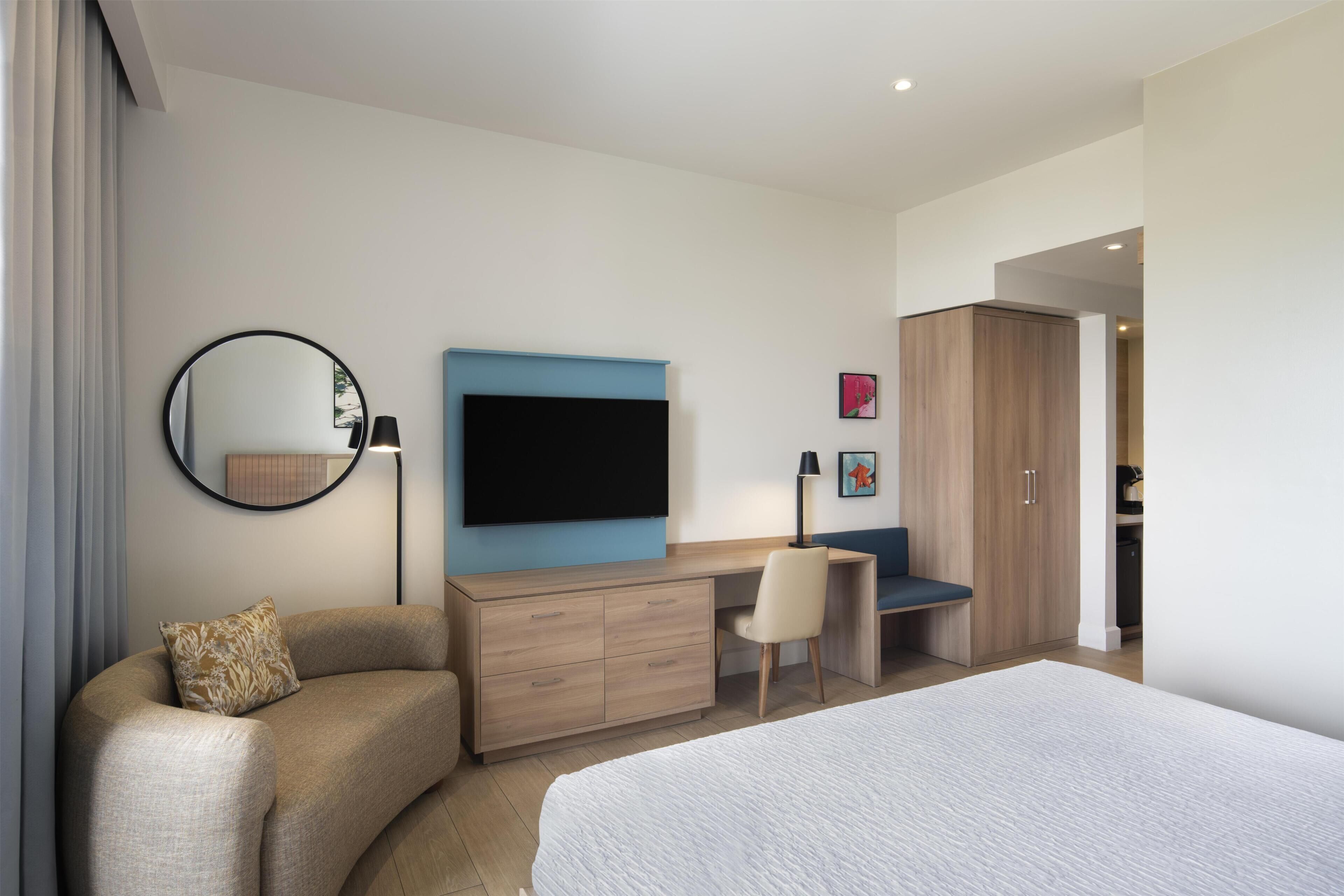 A modern hotel room with a large bed, a curved sofa, a desk with a chair, a flat-screen TV, a wardrobe, and a mirror.