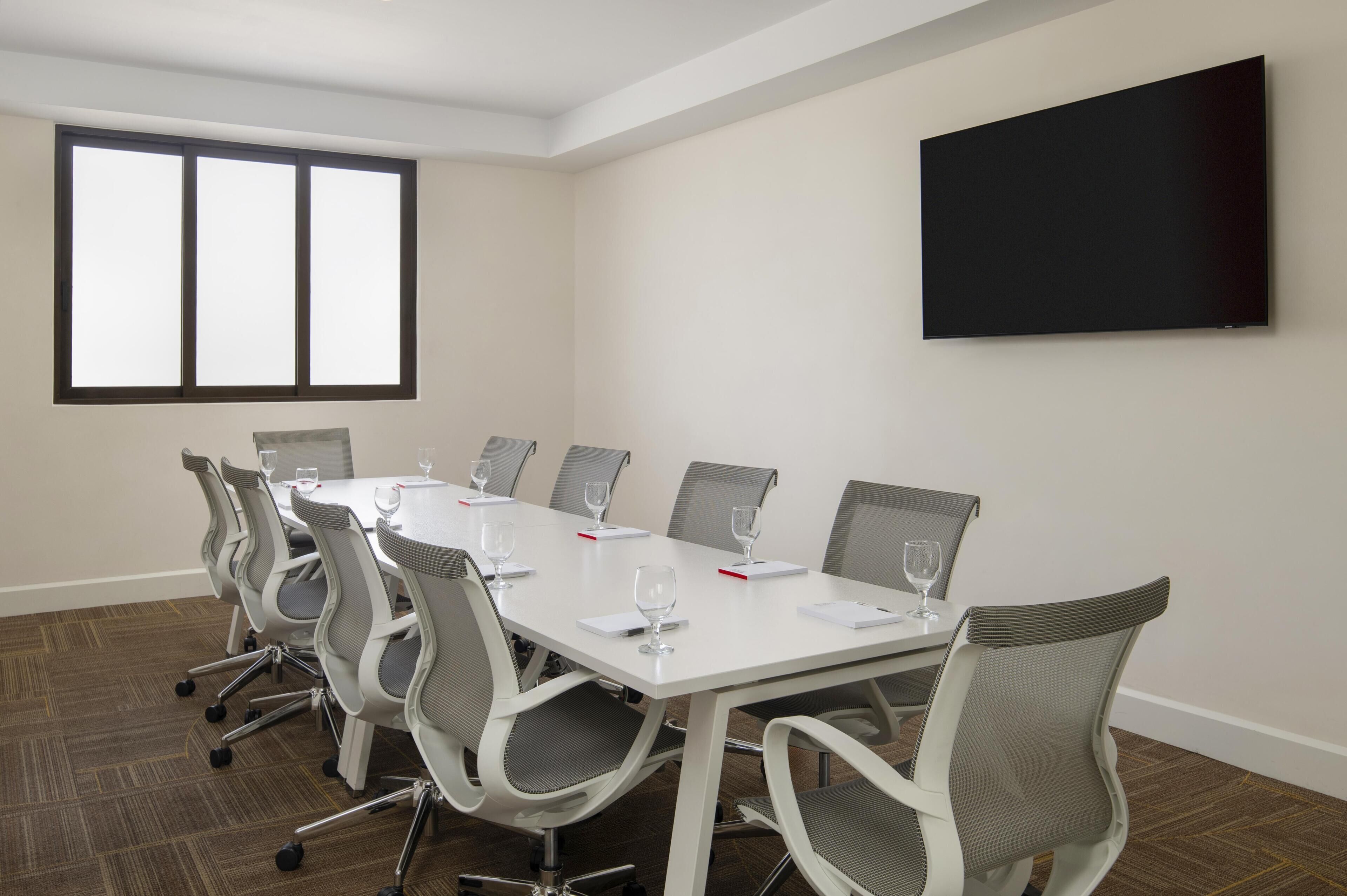 A conference room with a long table, chairs, a flat-screen TV, and a large window.