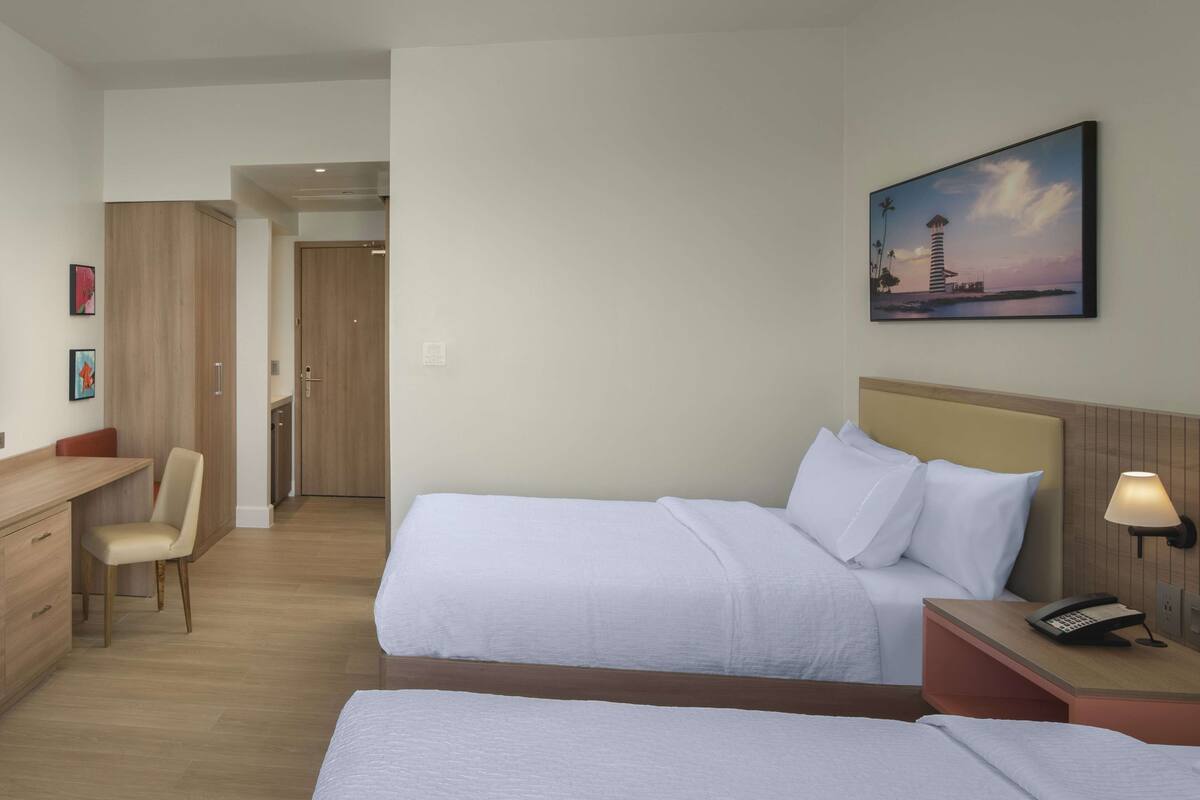 Une chambre d’hôtel avec deux lits, un bureau, une chaise, un tableau et une table de chevet avec une lampe.