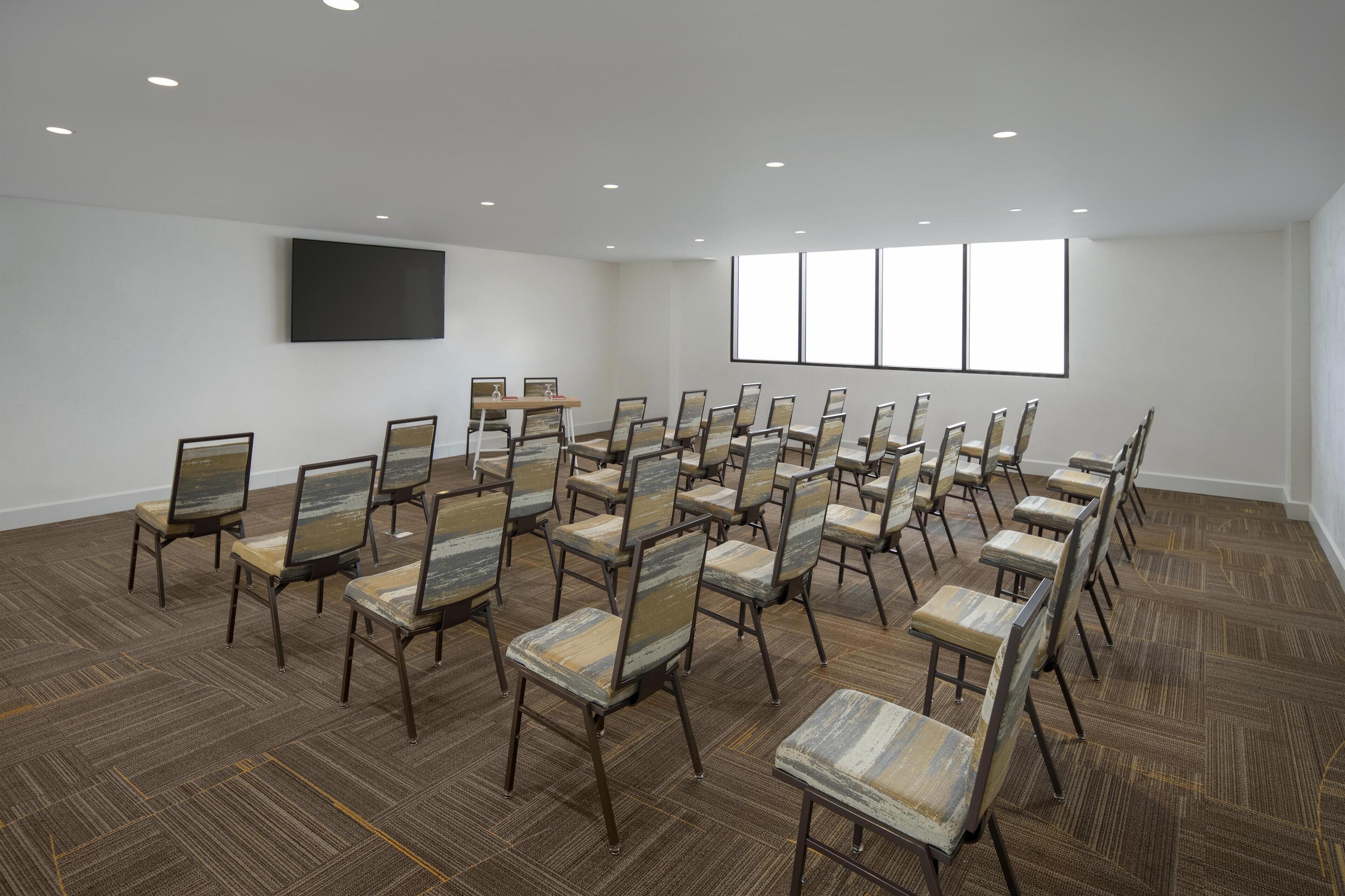 Une salle de conférence avec des rangées de chaises, un grand téléviseur à écran plat et une fenêtre équipée de stores.