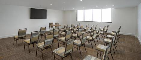 Une salle de conférence avec des rangées de chaises, un grand téléviseur à écran plat et une fenêtre équipée de stores.