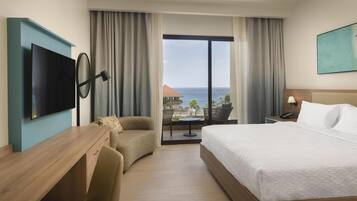 A hotel room with a bed, a desk, a TV, a chair, a table, and a view of the ocean.