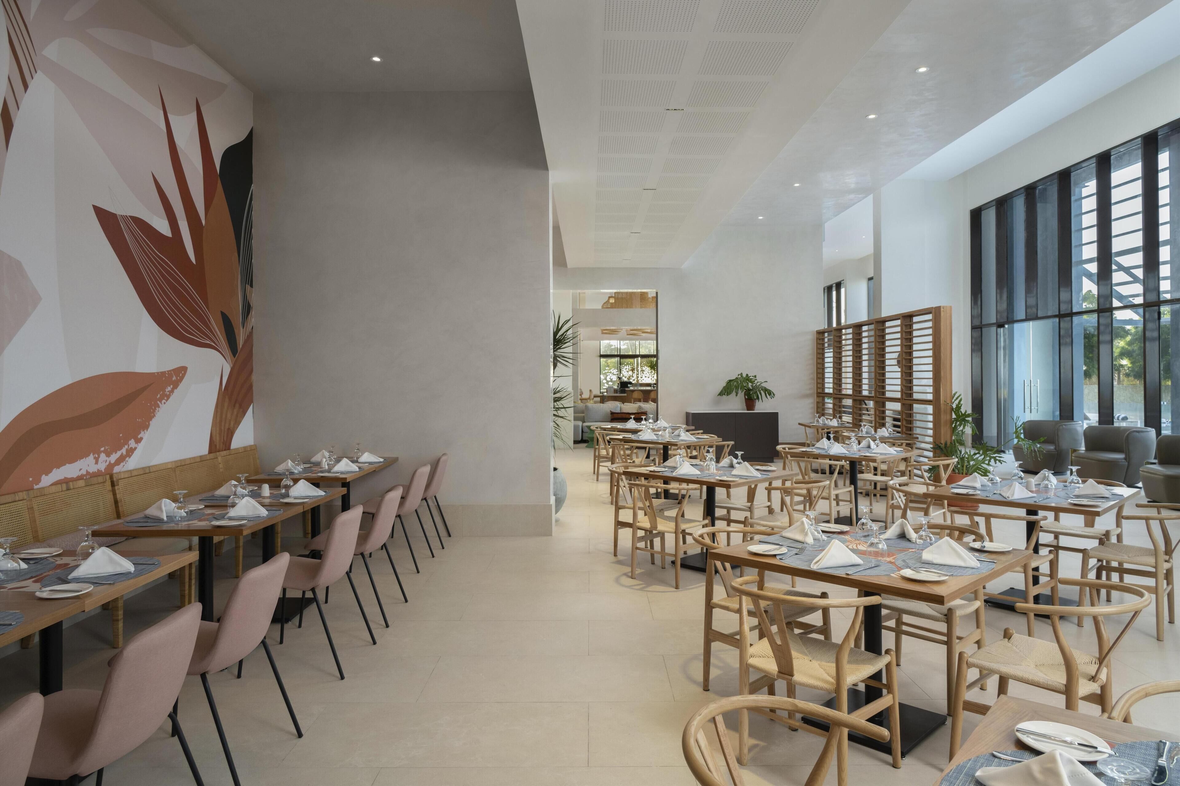 Un comedor moderno con mesas y sillas largas, un mural grande en la pared y ventanas amplias.