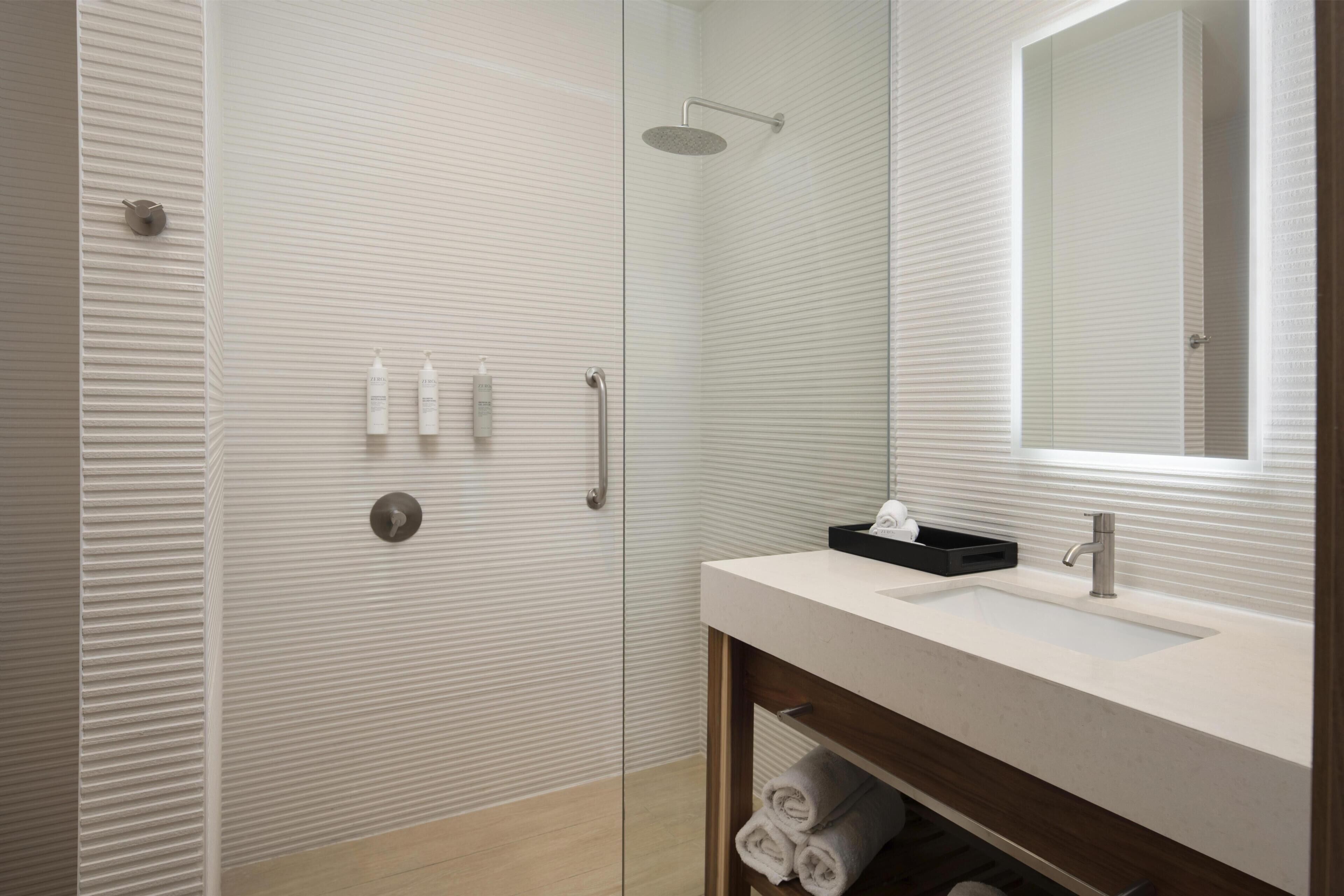 A modern bathroom with a glass shower enclosure, a white sink with a black soap dish, and a wooden vanity with towels.