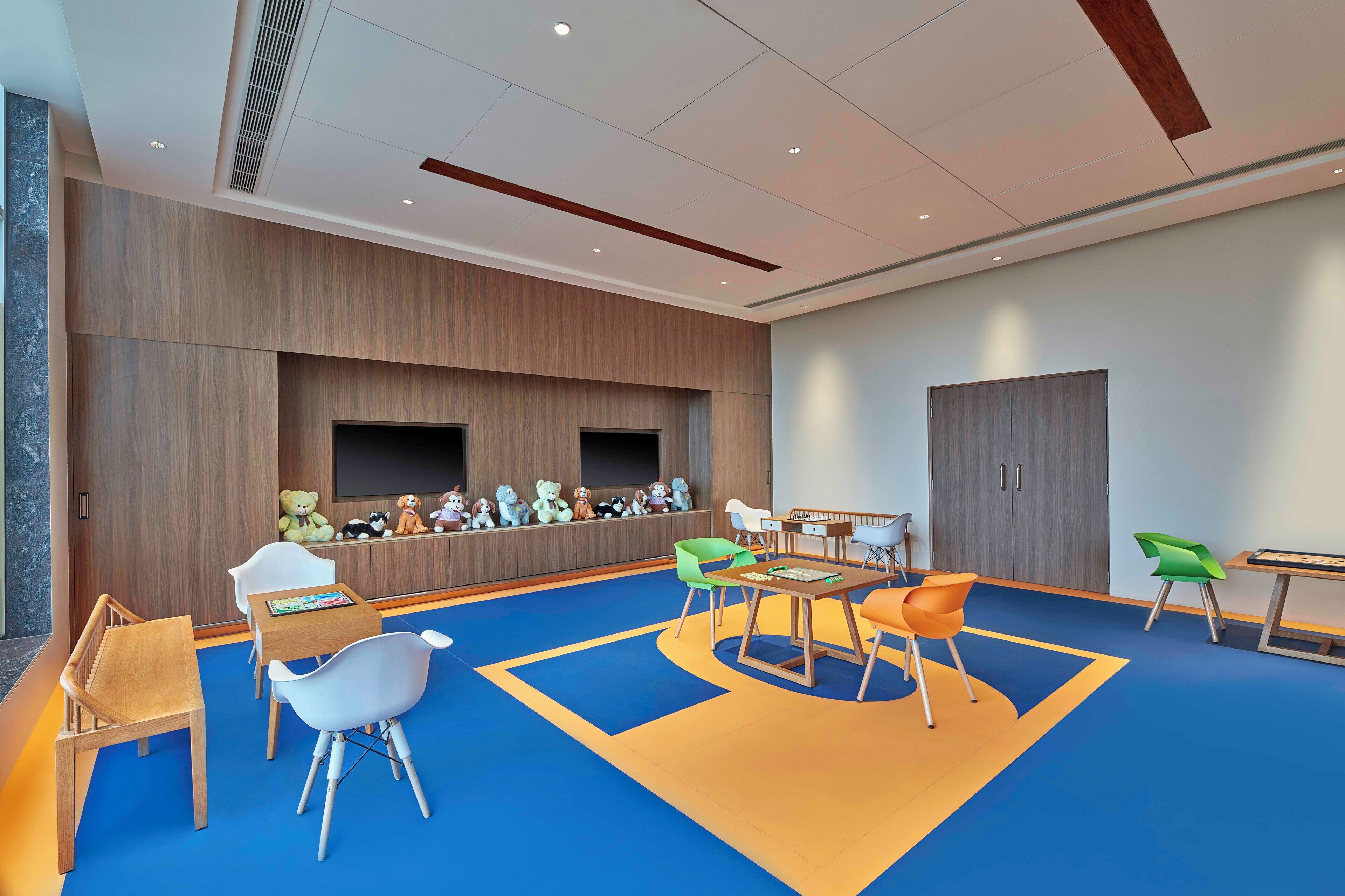 A children's playroom with a blue carpet, wooden play tables, and colorful chairs.