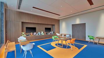 A children's playroom with a blue carpet, wooden play tables, and colorful chairs.