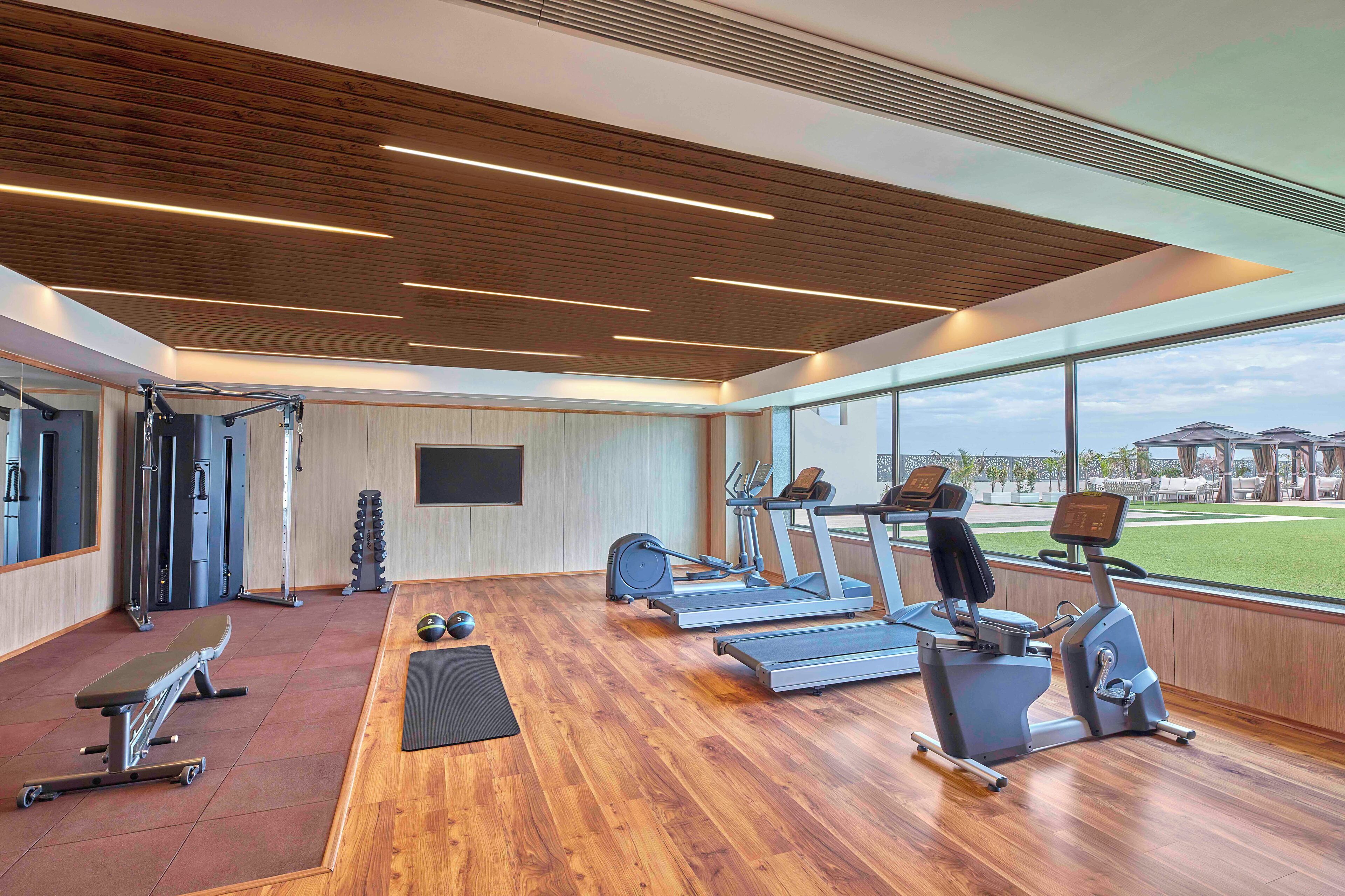 Un gimnasio moderno con suelo de madera, equipo de ejercicio y ventanas amplias con vistas al exterior.