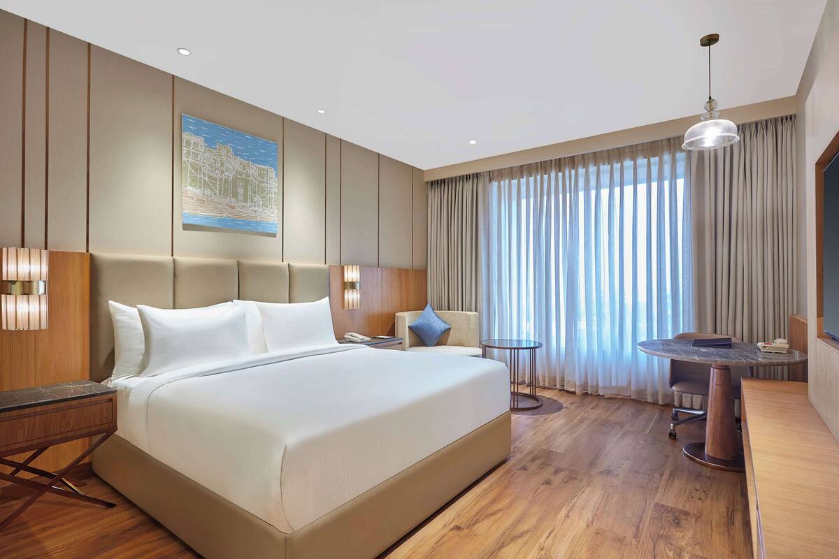 Una habitación de hotel moderna con una cama grande, una mesita pequeña, una silla, un escritorio y un ventanal con cortinas.