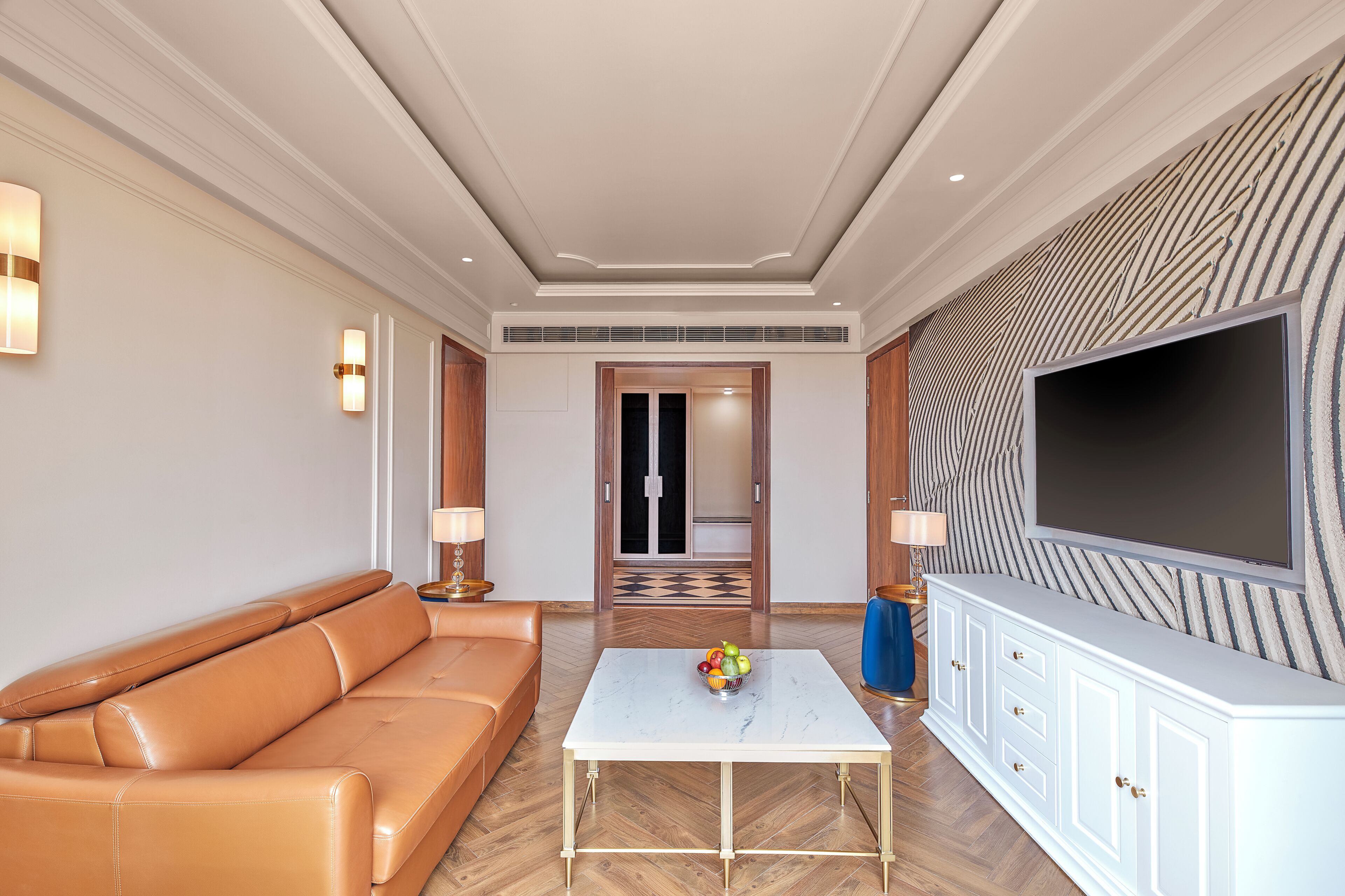 A modern hotel room with a leather sofa, a marble coffee table, a flat-screen TV, and a patterned wall.