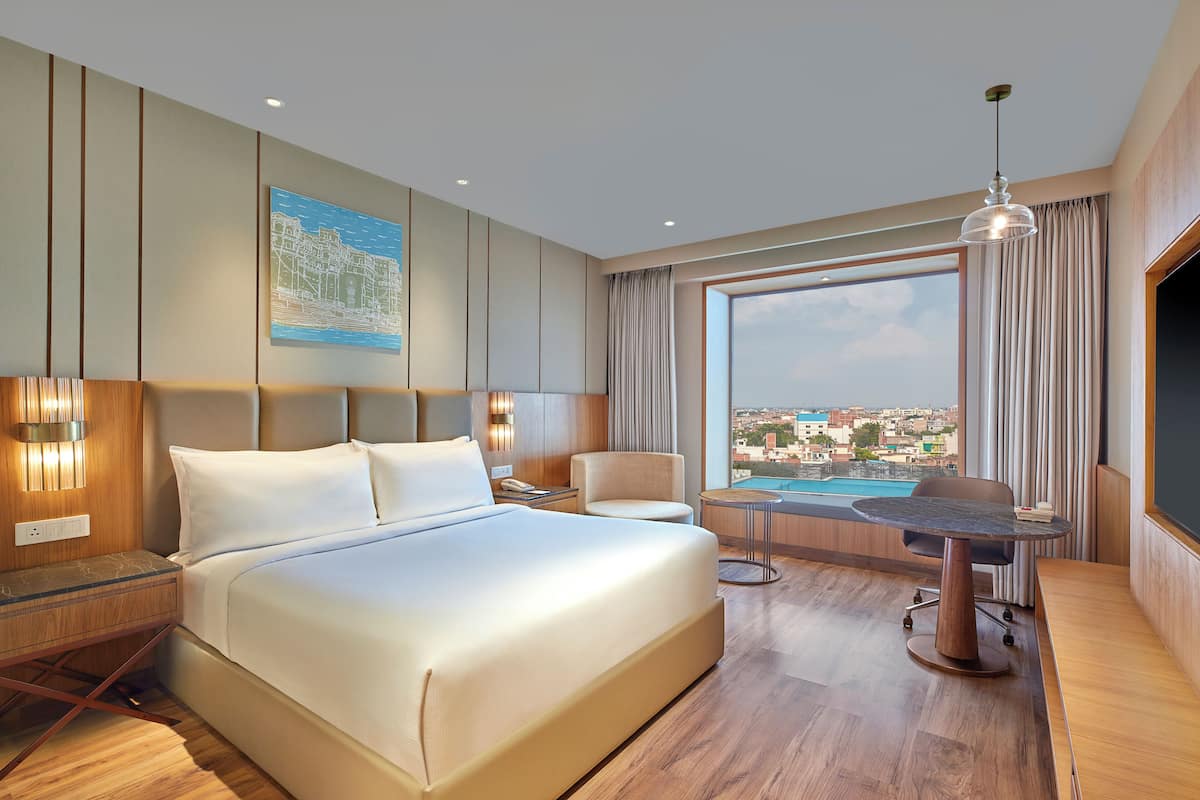 Habitación de hotel con una cama grande, un escritorio con silla, vistas a un paisaje urbano y una piscina.