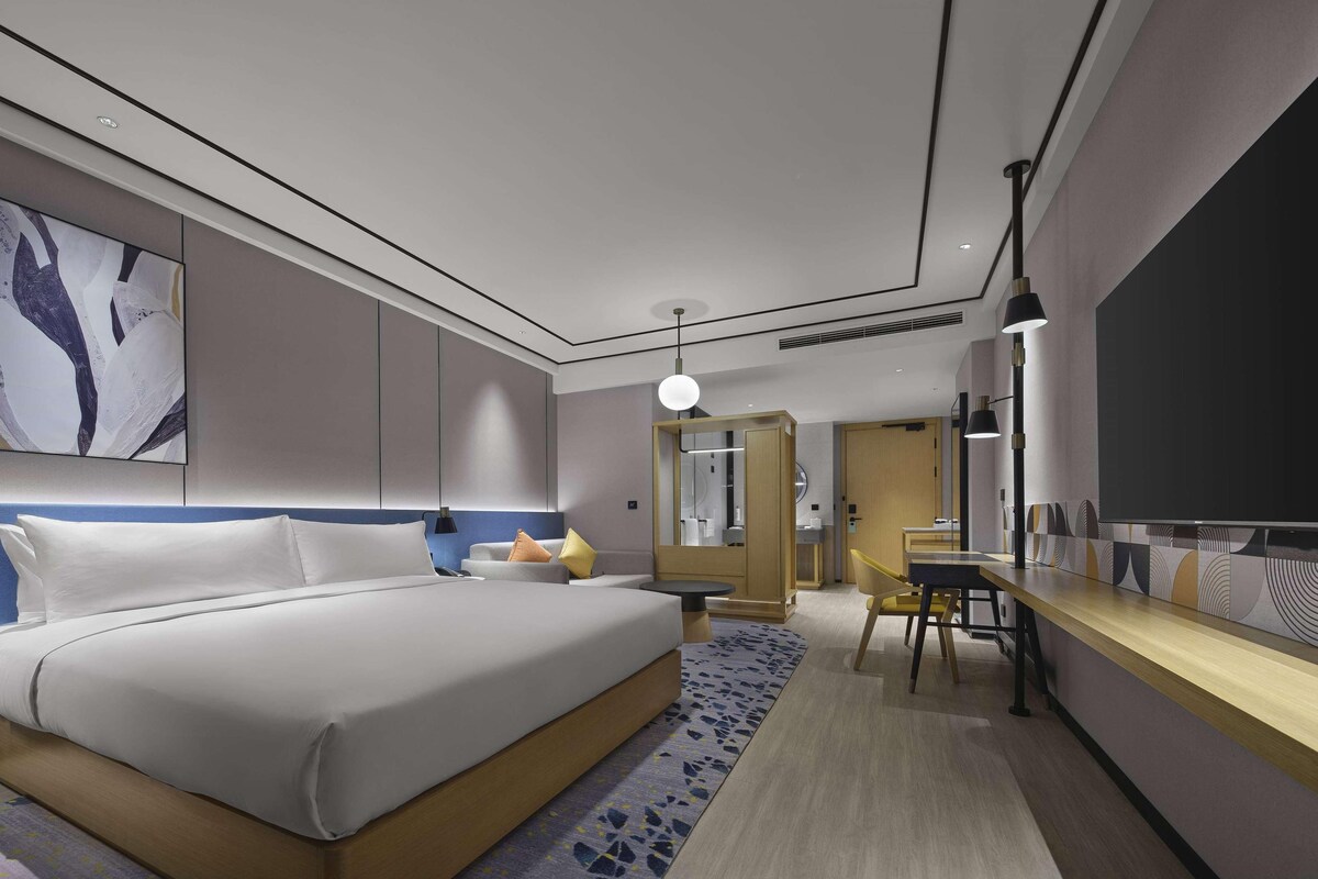 Une chambre d’hôtel moderne, dotée d’un grand lit, d’un bureau, d’une chaise, d’une télévision et d’une salle de bain visible à travers une porte ouverte.