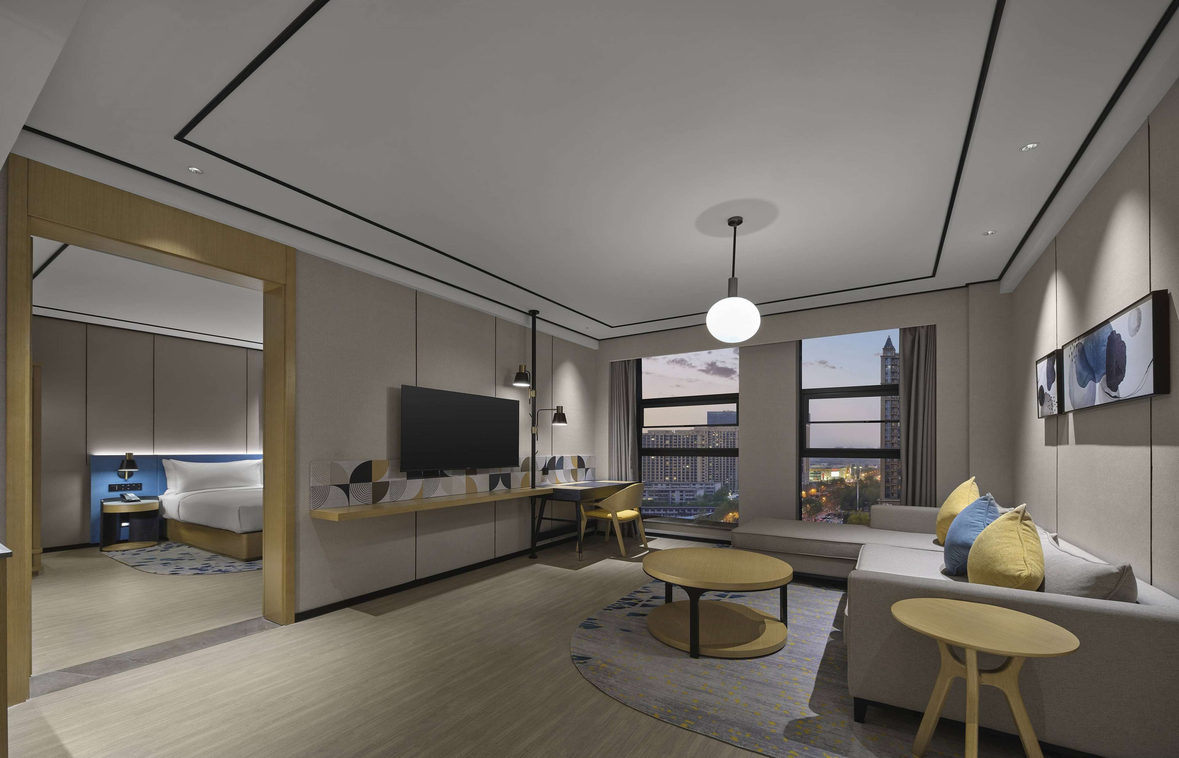 Una habitación de hotel moderna con una cama grande, televisión, escritorio y sofá. Se ve una ventana con vista a la ciudad.