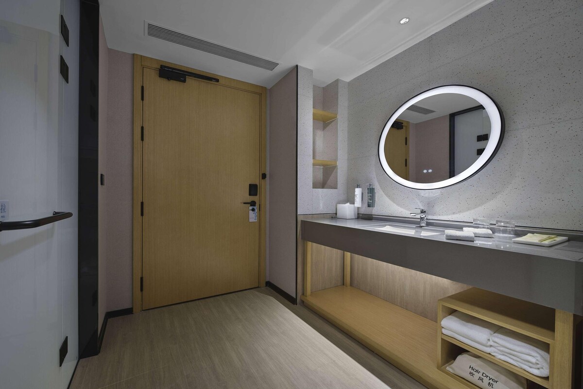 Un baño de hotel con una puerta de madera, un espejo redondo, un lavamanos y un perchero para toallas.