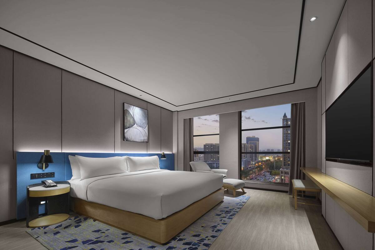 Habitación de hotel moderna con una cama grande, un escritorio, una silla, un televisor y vistas a la ciudad.