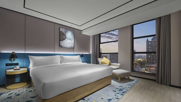 Una habitación de hotel moderna con una cama grande, una mesita de noche, una silla y vistas de la ciudad a través de amplios ventanales.
