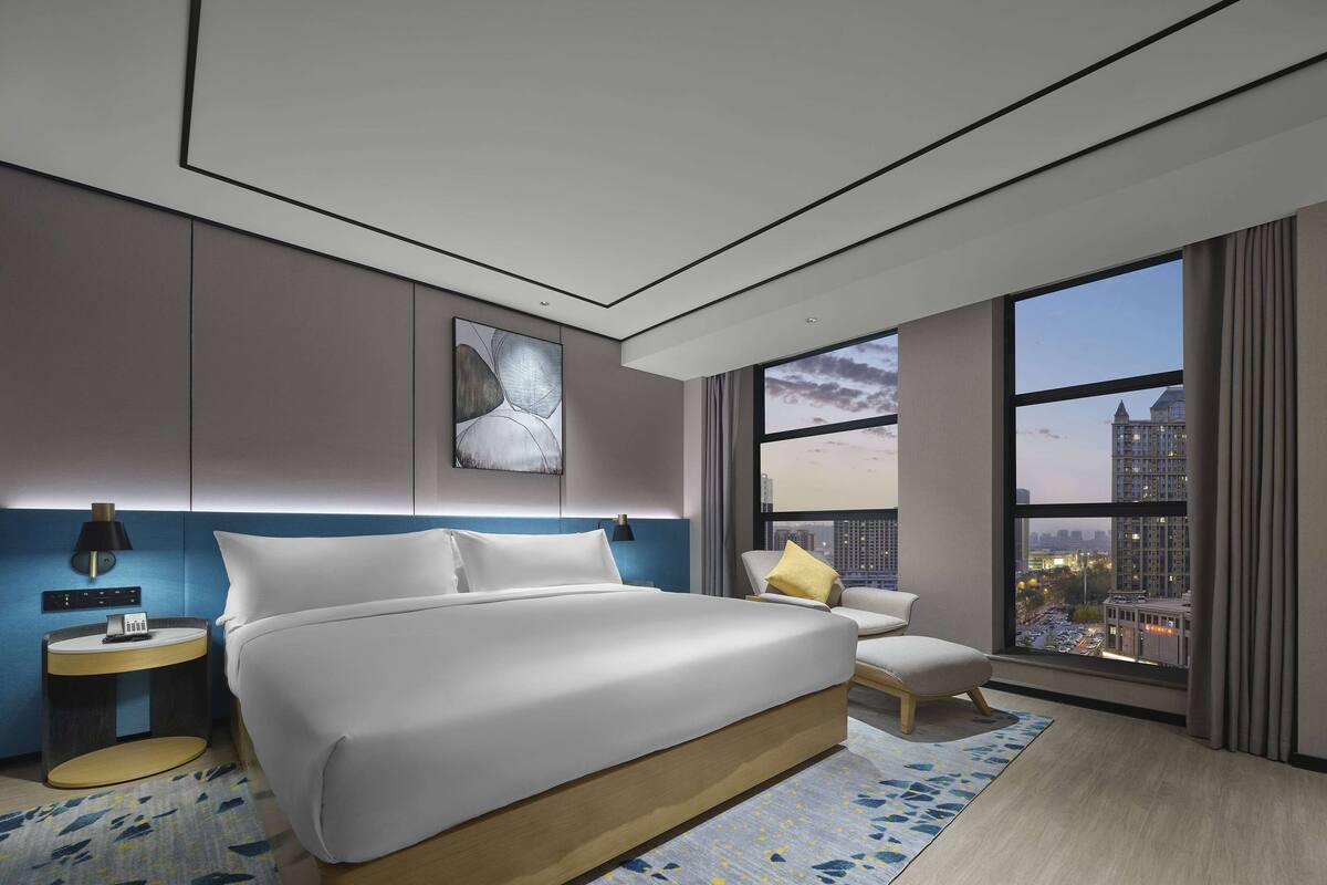 Una habitación de hotel moderna con una cama grande, una mesita de noche, una silla y vistas de la ciudad a través de amplios ventanales.