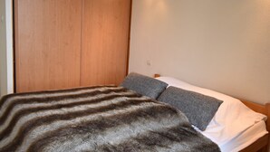 Une chambre avec une armoire en bois, un lit avec une literie grise et blanche, et un luminaire fixé au mur.