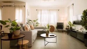 Ein modernes Wohnzimmer mit Sofa, Couchtisch, Sessel und TV-Möbel. Mehrere Blumentöpfe und eine Lampe sind ebenfalls vorhanden.