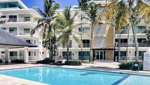 Ein Schwimmbecken mit Liegestühlen und Palmen, umgeben von modernen Gebäuden.