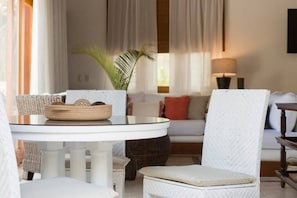 Um quarto com uma mesa redonda branca, cadeiras de vime, um sofá branco com almofadas vermelhas e um abajur.
