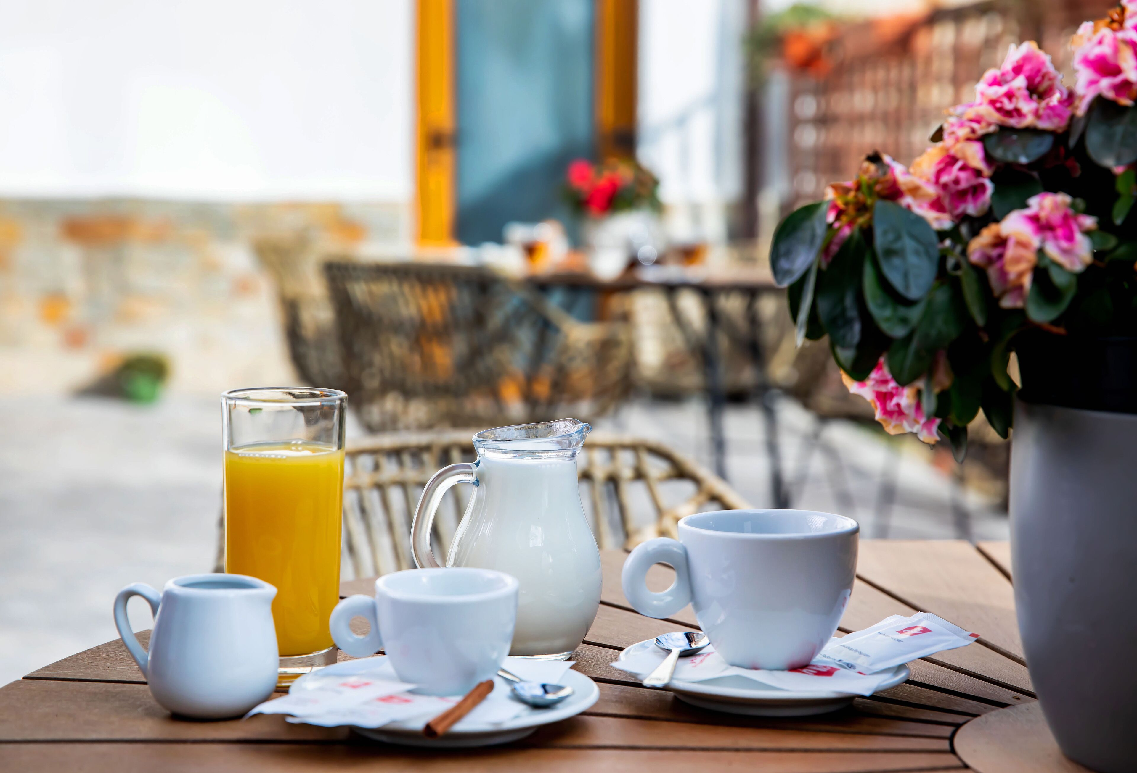 Un desayuno con una jarra de leche, un vaso de jugo de naranja y tazas blancas sobre una mesa de madera.