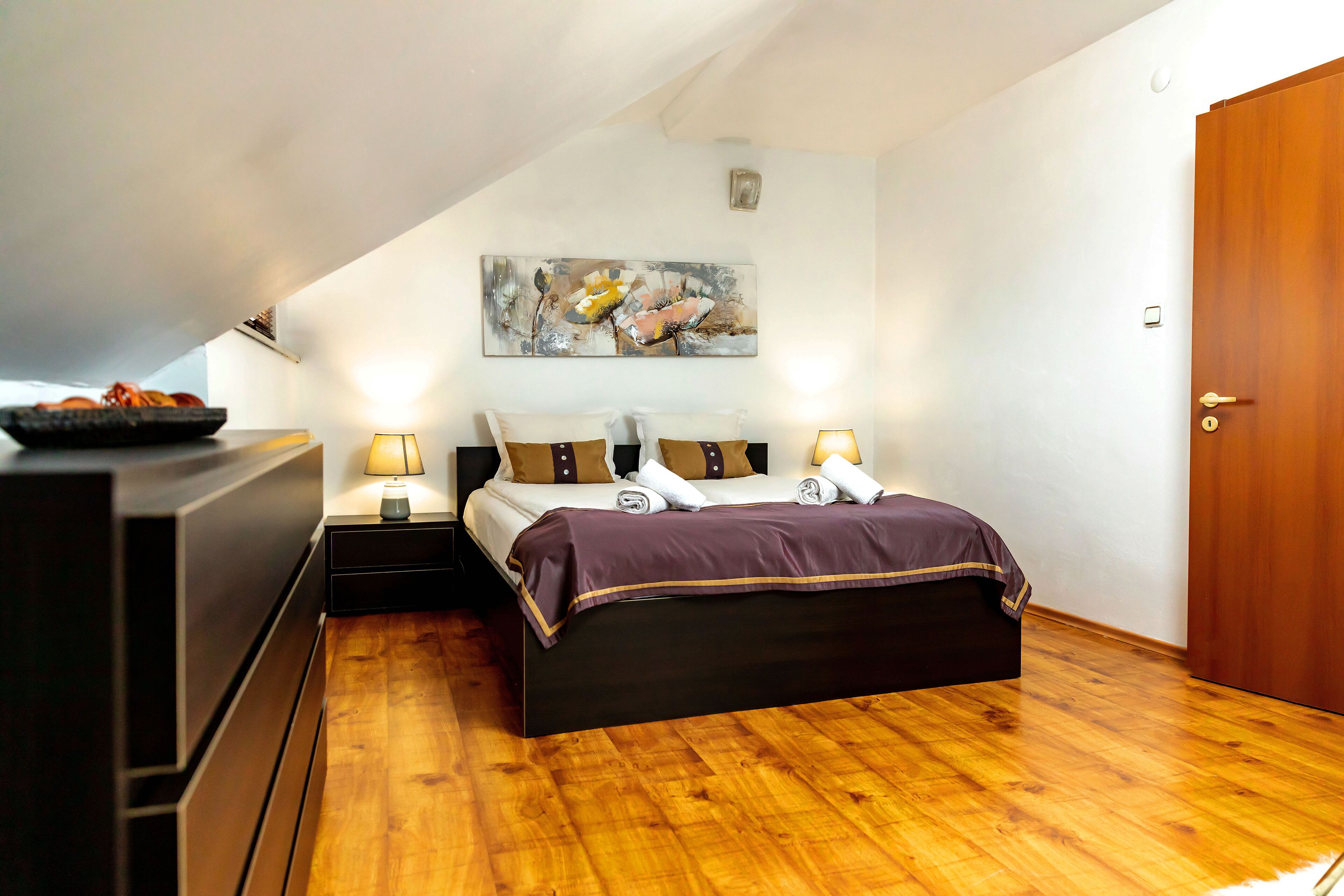 Un dormitorio con piso de madera, una cama con cubrecama morado, dos lámparas de noche y un cuadro en la pared.