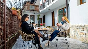 Dos personas sentadas en una mesa al aire libre con una bebida, en un patio con un edificio blanco y una valla de madera.