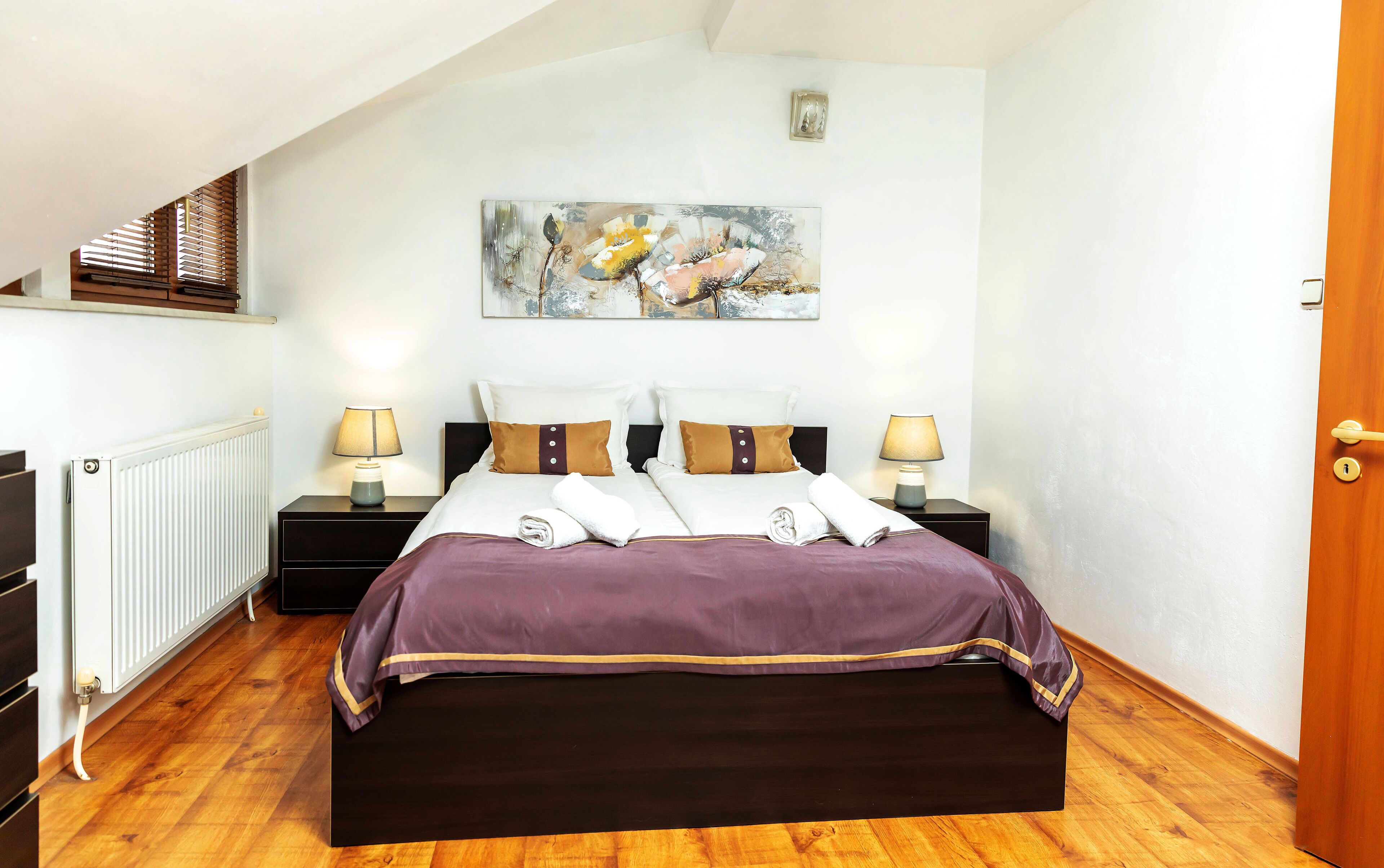 A neatly arranged bedroom with a large bed, two bedside tables with lamps, a radiator, and a painting on the wall.