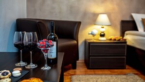 Une chambre d’hôtel chaleureuse, avec une table en bois mise en place pour un repas : on y trouve des verres à vin, une bouteille de vin et un dessert aux fruits rouges. Une lampe est posée sur une table de chevet, et on aperçoit un lit au fond de la pièce.