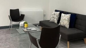 Ein modernes, minimalistisches Zimmer mit Glastisch, zwei schwarzen Stühlen, einer dunklen Couch mit gemusterten Kissen und Einbauleuchten.