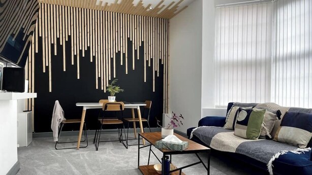 Ein modernes Wohnzimmer mit schwarzer Wand, Holzlatten, einem blauen Sofa und einem Essbereich mit Tisch und Stühlen.