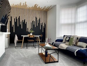A modern living room with a black wall, wooden slats, a blue sofa, and a dining area with a table and chairs. - Luxury 2 Bed Apt- Leicester City- Free Parking (Leicester)