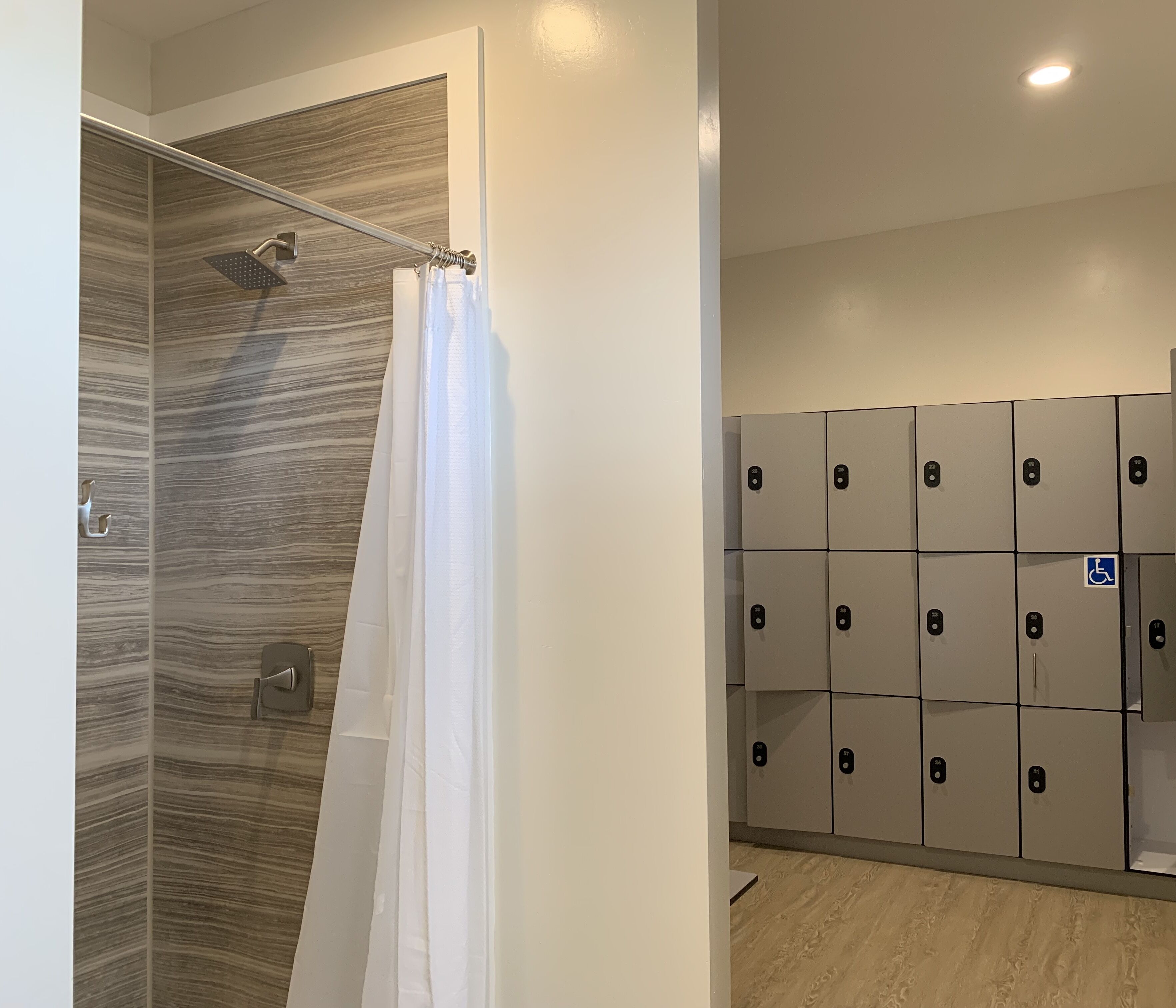 A shower with a wooden wall and a curtain, a row of lockers with a wheelchair accessibility sign, and a light-colored floor.